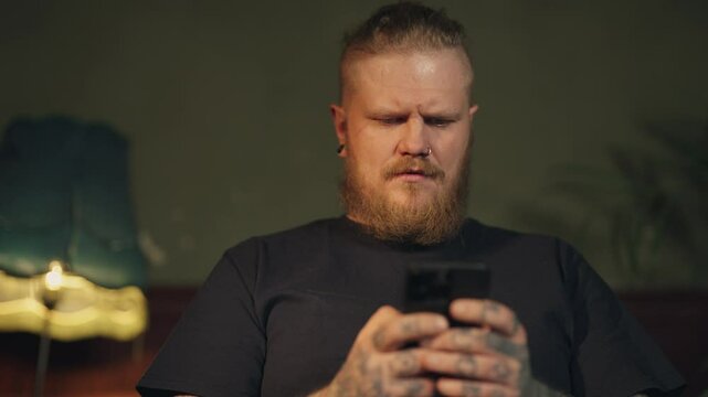 Male smartphone user surfing internet by mobile applications and browser at home . Portrait of adult man with beard, piercing and tattoo holding mobile phone in hands, online shopping and chatting