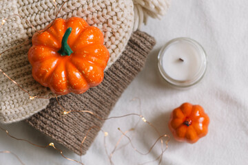 A candle in a glass jar on the background of a folded knitted sweater, and a beige hat with orange pumpkins lies on a white table in the room. Autumn decor. Top view