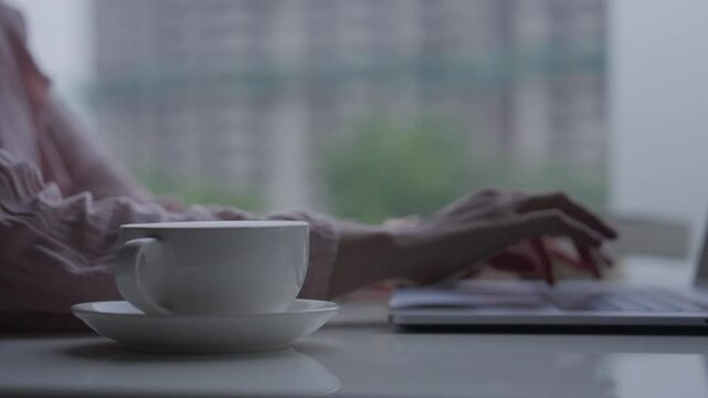 A high speed side close-up shot of an Indian female working and typing on a laptop, with a foreground of a white cup saucer kept on the table and just her hands visible while typing