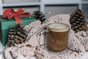 Winter still life. A glass cup of coffee with milk, big cones, lights, a green gift box with a red bow. Modern christmas rustic eco friendly decor, scandinavian winter hygge. Atmospheric christmas