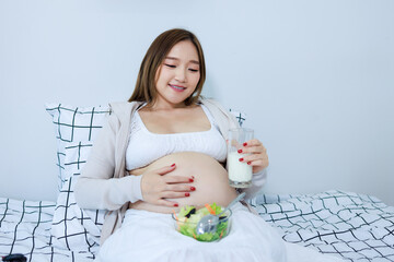 Pregnant asian woman drinking milk and salad on bed in bedroom at home