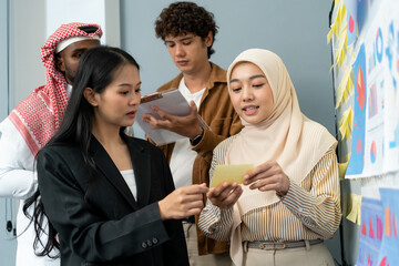 Muslim businesswoman in a hijab leading a presentation, pointing at a board, while a colleague takes notes during a team discussion.