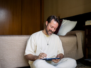 A man smiles while seated on the floor of a cozy room, focused on sketching with a stylus on a...
