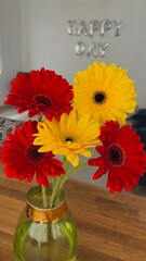 a bouquet of red and yellow gerberas in a glass vase on the background of a wall with the inscription happy day