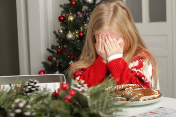 Sad, frustrated little girl in Christmas outfit and Santa hat sitting at the table with gingerbread cookies, winter holiday stress concept.	
