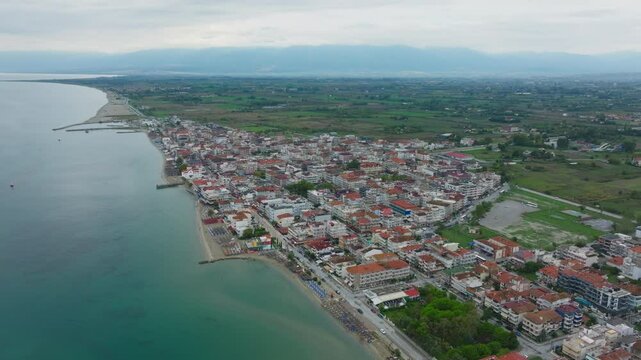 The camera is panning over Paralia Katerinis, a popular seaside resort town on the Aegean Sea, showcasing its long sandy beach and bustling promenade