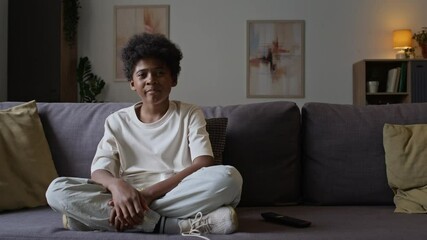 Full portrait of smiling little African American boy sitting with crossed legs on sofa and eating popcorn while watching TV at home, spending free time, looking at camera
