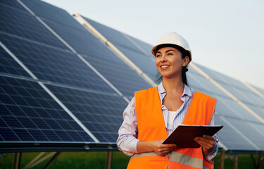 Black notepad in hands. Female worker engineer in uniform is near solar panels