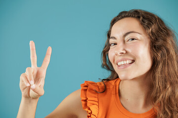 Portrait of a young woman on a blue background. She poses waving with her hand, showing a v with her fingers. Empowered and independent woman.
