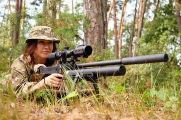A skilled marksman in camouflage gear carefully aims with a sniper rifle while lying in a forested...