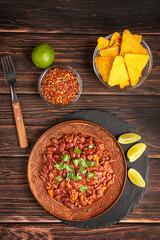 Flat lay with homemade spicy Mexican chili con carne stew made of pepper flakes, minced beef meat, tomatoes, and kidney beans on craft plate with lime slices and tortilla chips on dark wooden table