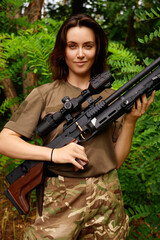 A young woman poses confidently in military attire with a sniper rifle surrounded by dense green foliage during daylight