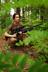 A soldier in tactical gear crouching in a lush green forest, holding a rifle and scanning the...