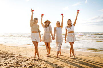 Happy women with glasses of wine walking on the beach. Celebrating bachelorette party, drinking wine at sunset. Holidays, nature, relax concept.