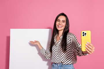 Cheerful young woman taking a selfie with a yellow phone against a pink background, wearing casual...