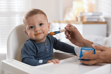 Mother feeding her cute little baby in high chair at home, closeup