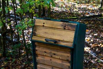 Close-up of a rustic compost bin in a woodland setting. The bin is dark teal with light brown planks, showing color variations and a reclaimed look. A metal handle is on the angled lid. 