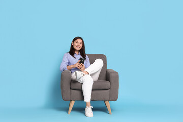 Smiling woman with smartphone resting in armchair on light blue background