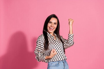 Excited young woman in striped shirt celebrates success against pink background in casual style