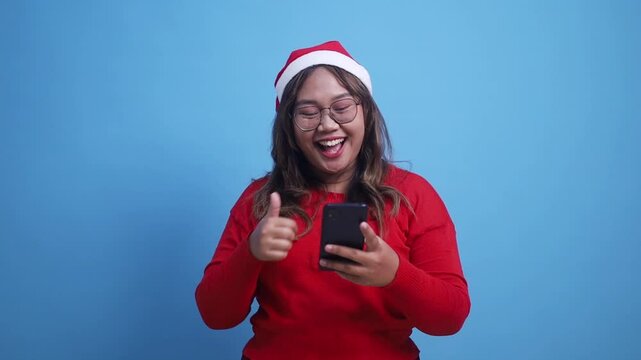 Young happy excited joyful Asian woman in Christmas clothes hold use mobile cell phone typing say wow yes just found out great big win news do winner gesture isolated on blue background