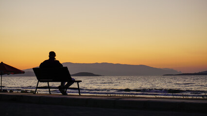 Person sitting alone on a seaside bench at sunset, surrounded by colorful umbrellas and mountains,...