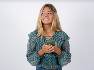 caucasian teen girl wearing green dress over white holding in hands mobile phone and looking aside with dreamy look.