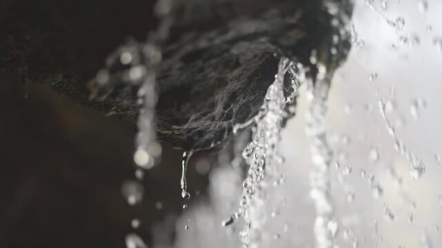 Clear droplets drip from rocky ledges in slow motion, each one distinct. Behind, a spring waterfall flows, representing peace and water scarcity issues.
