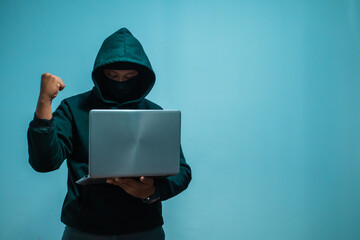 Faceless anonymous hacker in black hoodie and covering his face holding laptop with fists in joy and on dark background, Financial data theft concept, Empty space for text, copy space