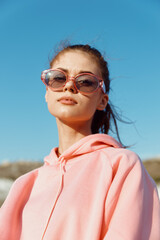 Stylish young woman relaxing on sandy beach in pink hoodie and sunglasses during sunny day