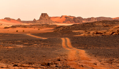 A road through the Sahara desert, ruts between the rocks, a winding road, orange sand and mountains all around.