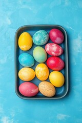 A tray of colorful eggs is displayed on a blue background. The eggs are arranged in a grid pattern, with some eggs being larger than others. The colors of the eggs include yellow, green, and red