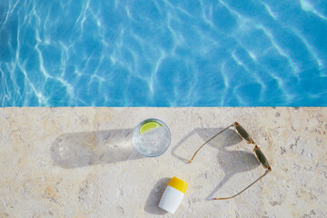 Overhead view of sunglasses, sun cream and drink by pool