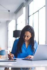 Long haired black businesswoman attentively writing at table with laptop