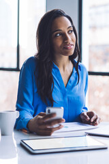 Thoughtful ethnic businesswoman using smartphone while sitting in office with devices