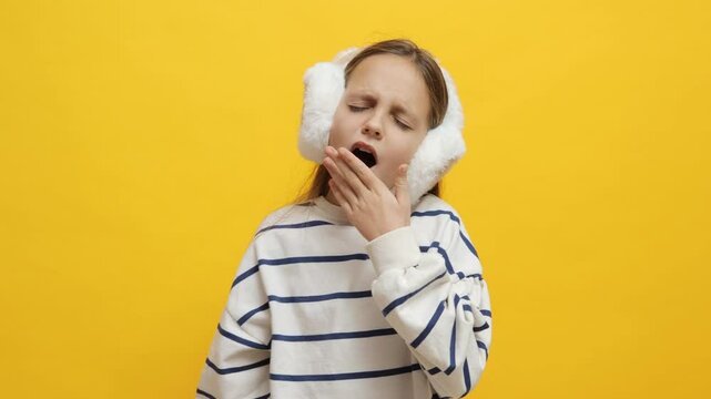 Little girl in fur headphones yawning feeling sleepy and tired showing exhaustion and drowsiness feeling boredom and fatigue waking up in early morning isolated on yellow background