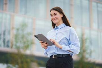 Young Woman in a Blue Shirt Uses a Tablet Outdoors in an Urban Environment During Daylight