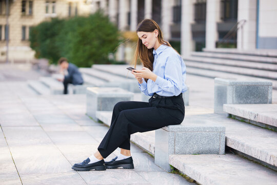 Young Woman Sitting on Steps in a Cityscape, Focused on Her Smartphone While a Man is in the Background
