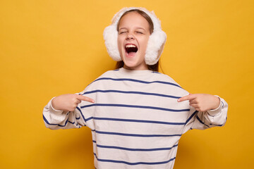 Excited little girl with fur headphones pointing at herself proudly screaming with happiness showing victory and success feeling self-important isolated on yellow background