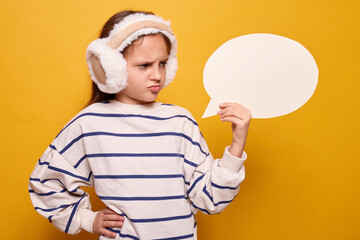 Sad little girl with fur headphones holding speech bubble with space for text looking disappointed at informing blank space for idea mockup isolated over yellow background