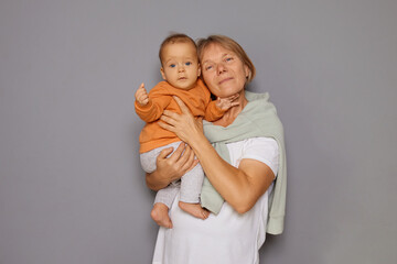 Cheerful female pensioner with joyful smile looking after her baby playing and caring for child isolated over gray background looking at camera with satisfied face