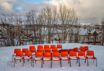 A cluster of orange chairs stands out against the snowy ground, surrounded by trees and a calm...