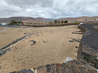 Plage de La Garita &agrave; Arrieta entour&eacute;e de murs de pierre volcanique, avec des traces de roches dispers&eacute;es et des montagnes arides en arri&egrave;re-plan sous un ciel nuageux.