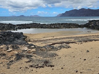 Une plage de sable avec des formations rocheuses volcaniques et des flaques d'eau, face à l'océan et encadrée par des montagnes, avec des îles visibles au loin sous un ciel nuageux.