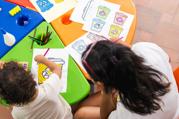 Top view of a Latina special education teacher guiding her student in an art project about recycling