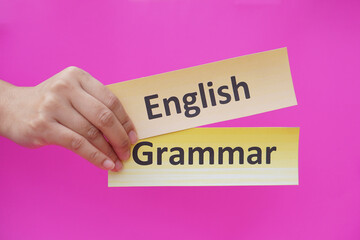Close up hands hold  word card paper with text English grammar. Pink background. Concept, English language grammar teaching lesson. Education materials.    