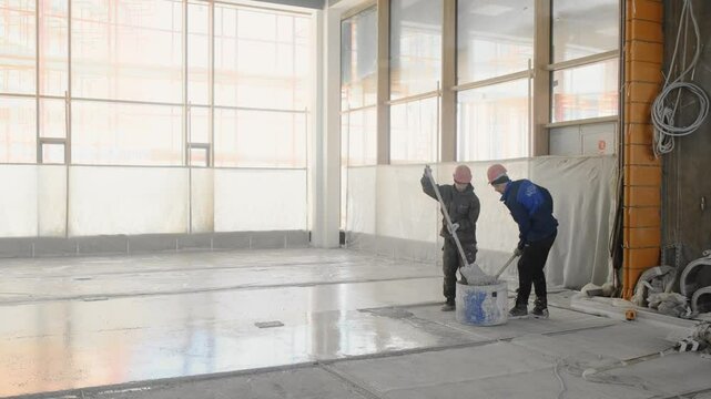 Two male workers in construction uniforms and with a helmet on their heads are leveling the concrete mixture on the floor with shovels. Concrete floor screed.