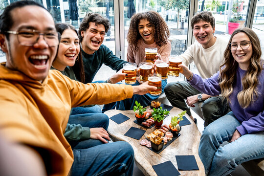 Happy multi racial hipster friends drinking and toasting beer at brewery pub - Group of young smile people taking selfie sitting at bar restaurant table - Food and beverage life style concept