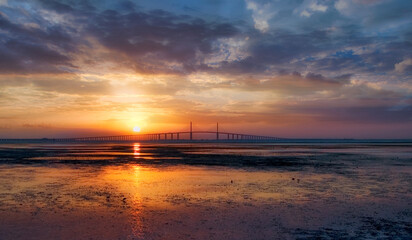 The sun rises behind the Sunshine Skyway Bridge, illuminating the shores of Fort DeSoto at low tide. It lights up the clouds and reflects off the water while shorebirds feed in the flats. Florida