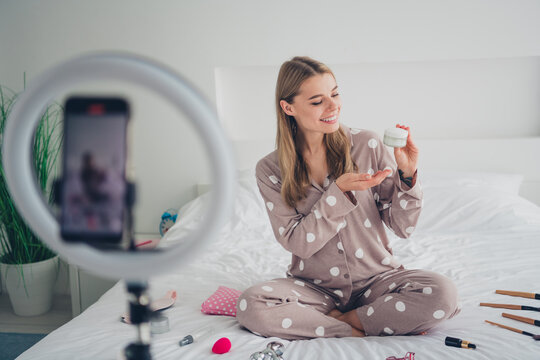 Young woman in polka dot pajamas sitting on bed recording a beauty vlog showcasing skincare in a bright bedroom
