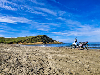Fototapeta premium A motorcycle standing alone on the shore of the sea on the beach of Zvernec in Albania, in the foreground are tire tracks in the sand, in the background is the sea surface and rocks with trees and bus